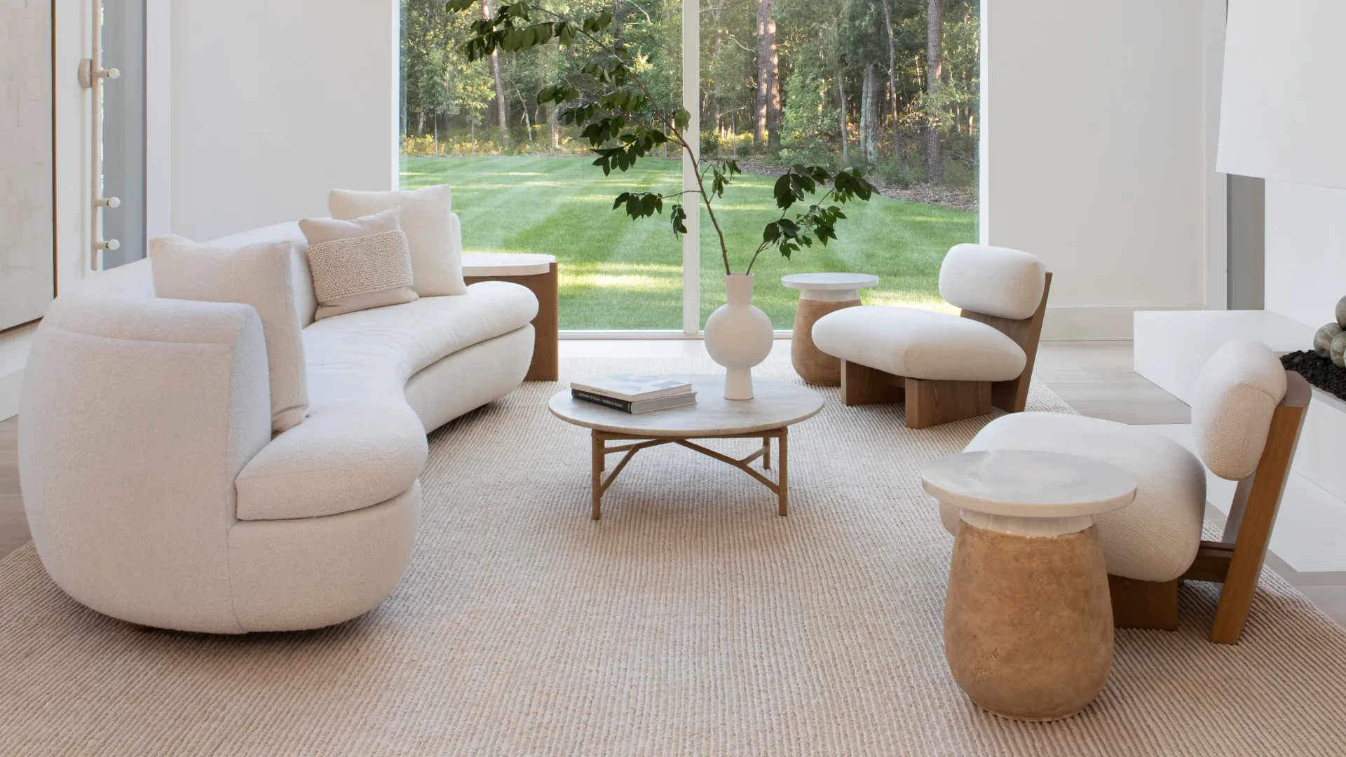 A curved cream boucle sofa and low oak lounge chairs around a round stone coffee table on a jute rug, facing a full height window onto a lawn.
