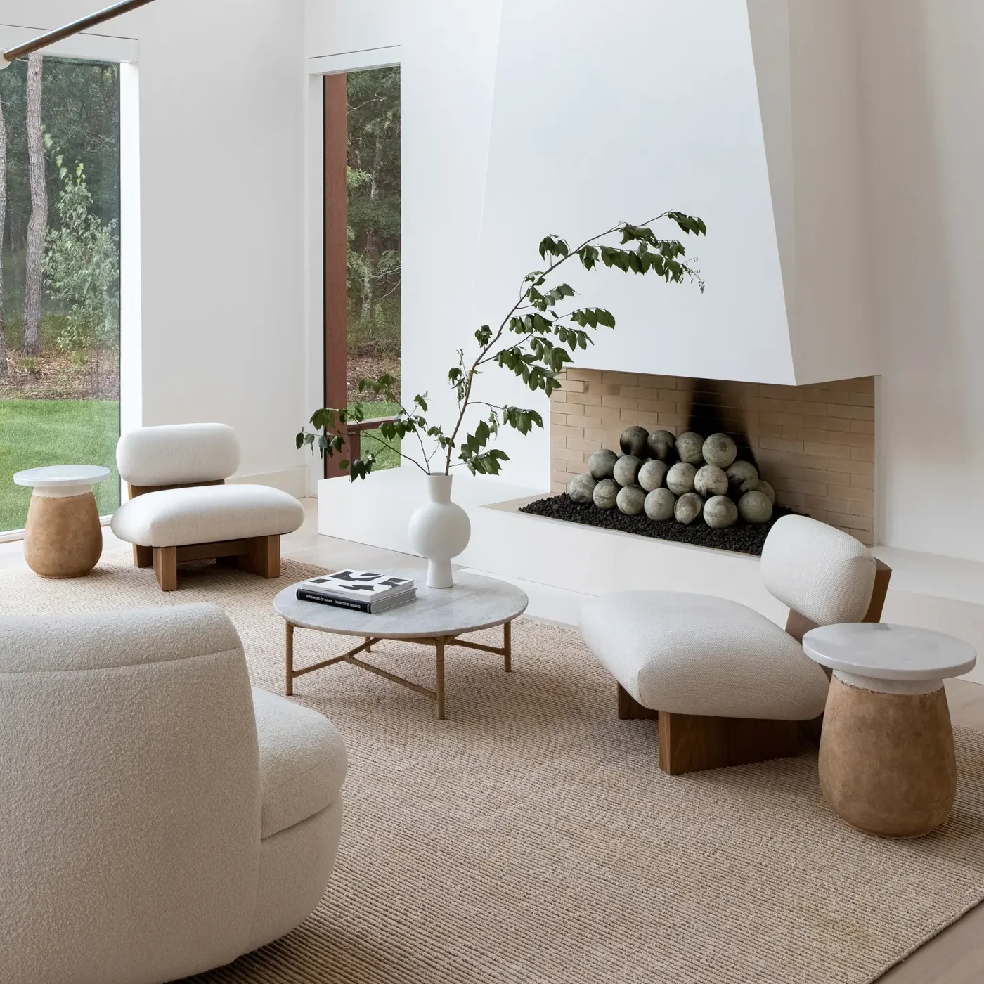 Low cream lounge chairs and a curved sofa around a round stone coffee table on a jute rug, in front of a white plaster fireplace.