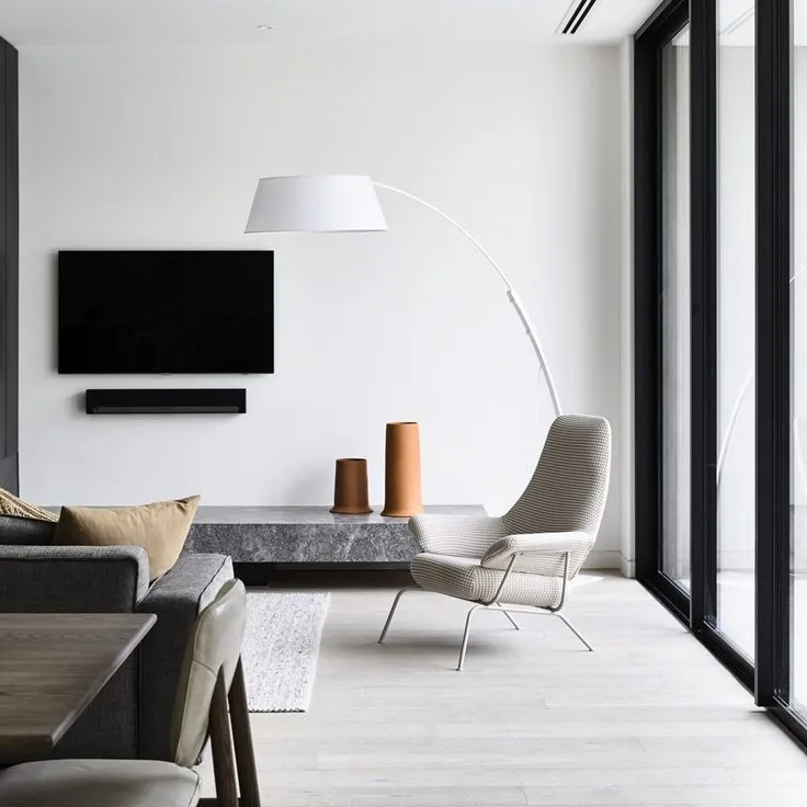 A grey sofa and a woven lounge chair beside a low stone bench holding two terracotta vessels, lit by a white arc floor lamp.