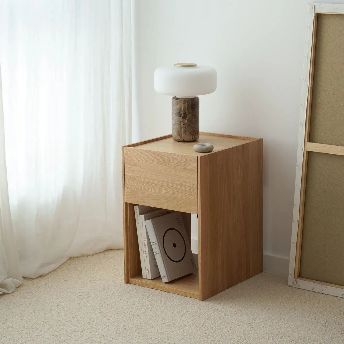 A small oak bedside table with a handleless drawer over an open shelf of books, beside a sheer linen curtain.