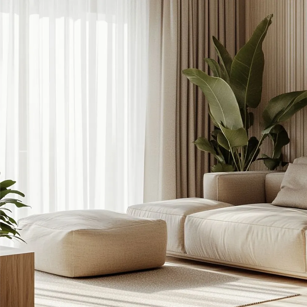 Low beige linen sofa and a floor cushion beside sheer curtains and a large green plant in a Japandi living room.
