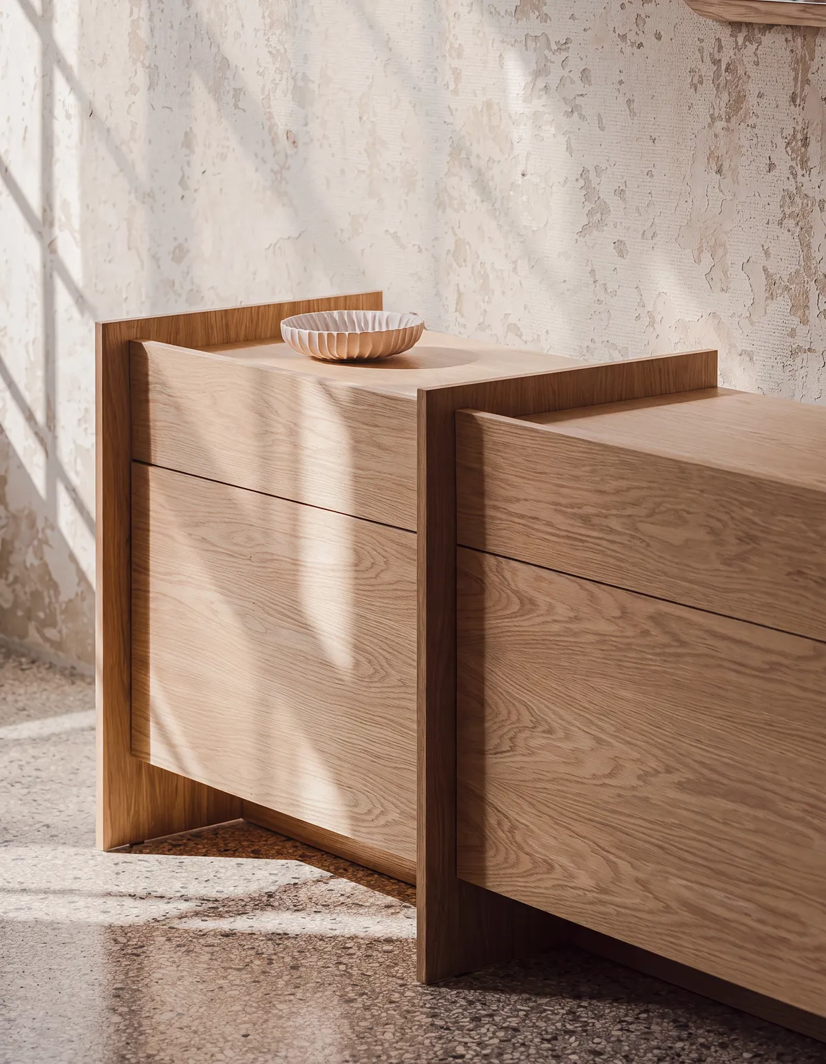Low pale oak storage unit with a fluted ceramic bowl on top, standing against a rough plaster wall.