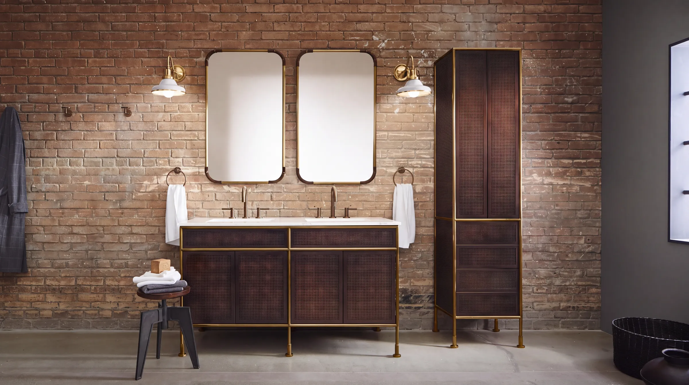 Industrial modern bathroom with a weathered brick wall, a dark wood double vanity and tall cabinet framed in brass, two brass-edged mirrors and white factory-style wall lights