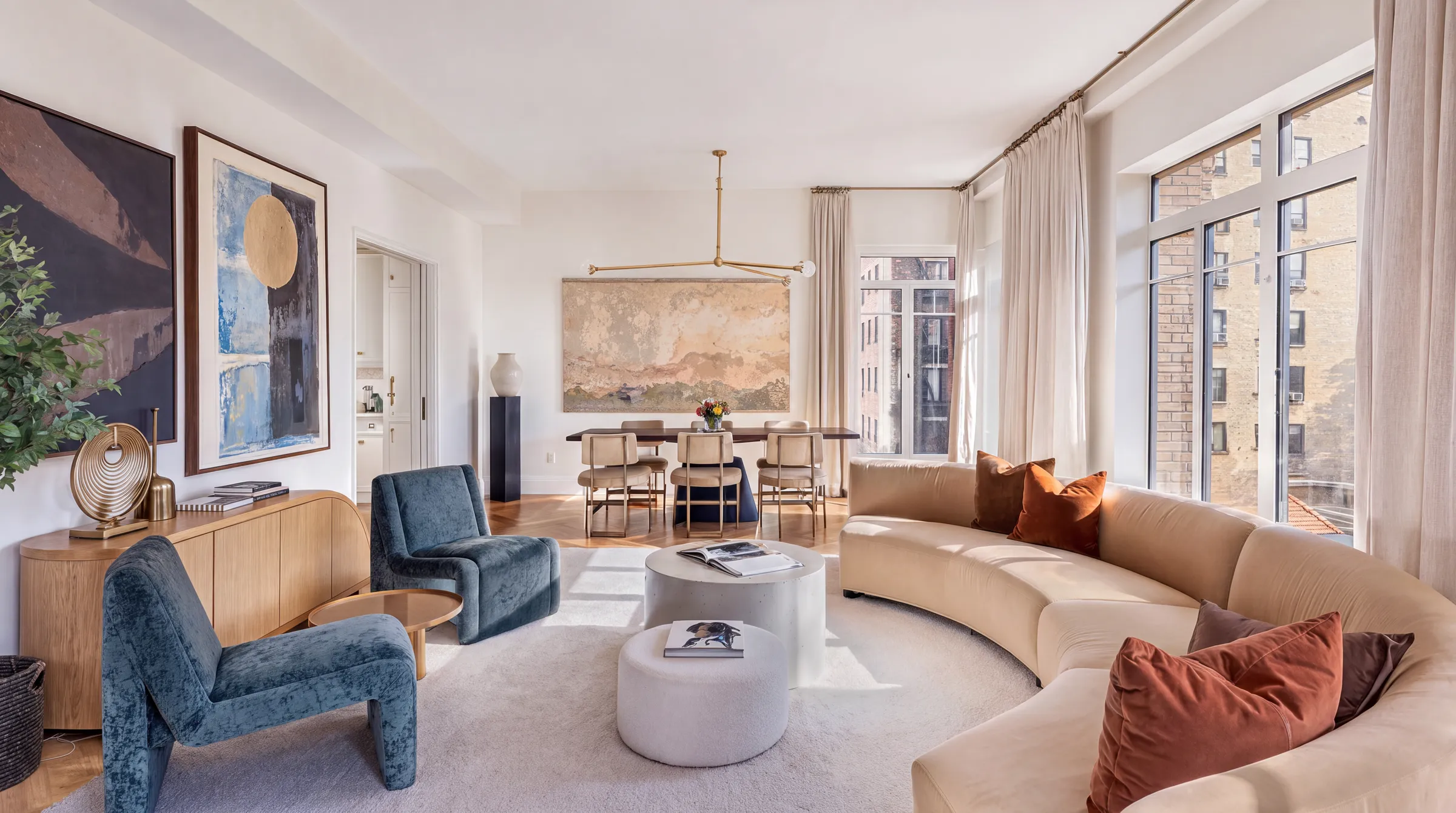 An organic modern living room with a curved cream sofa, two blue lounge chairs, rust cushions, a pale oak sideboard and round plaster tables under a brass pendant light.