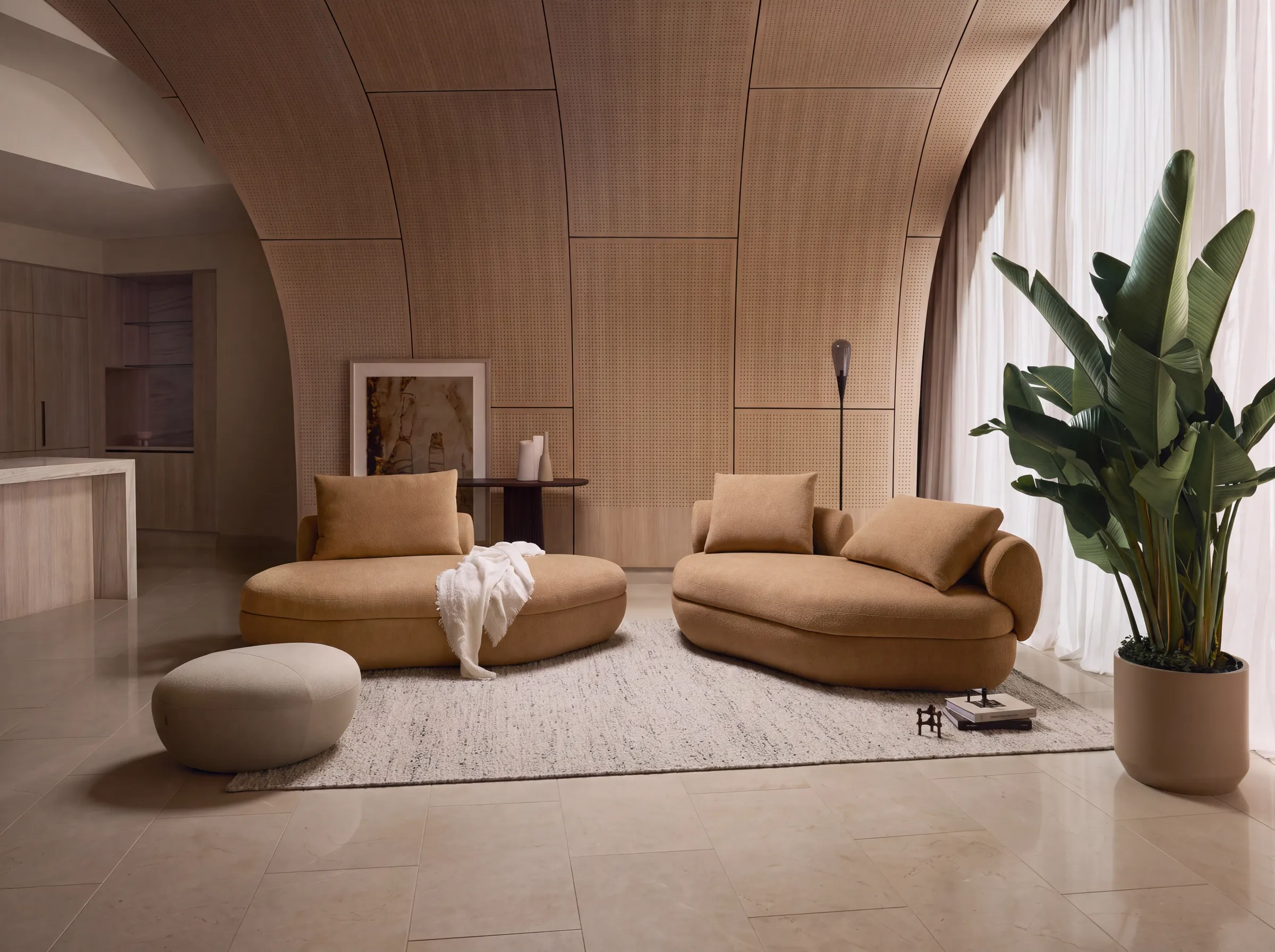 A living room with a curved perforated wood panel wall, two rounded camel sofas, a pebble shaped ottoman, a cream rug and a tall plant in front of sheer curtains.