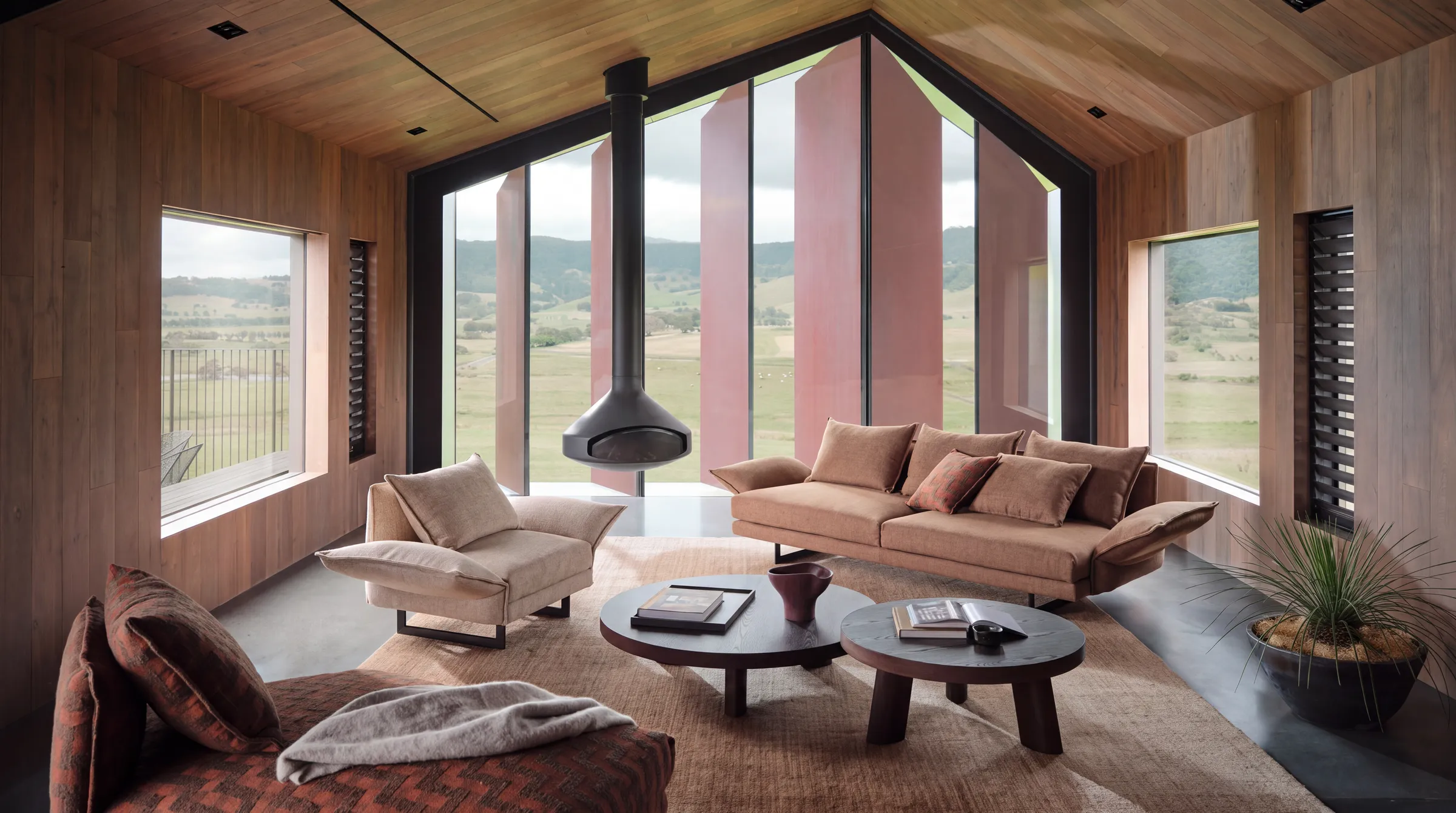 A living room lined in warm timber with a black suspended fireplace, a tan sofa, a rust armchair and two round dark wood coffee tables in front of large windows facing green hills.