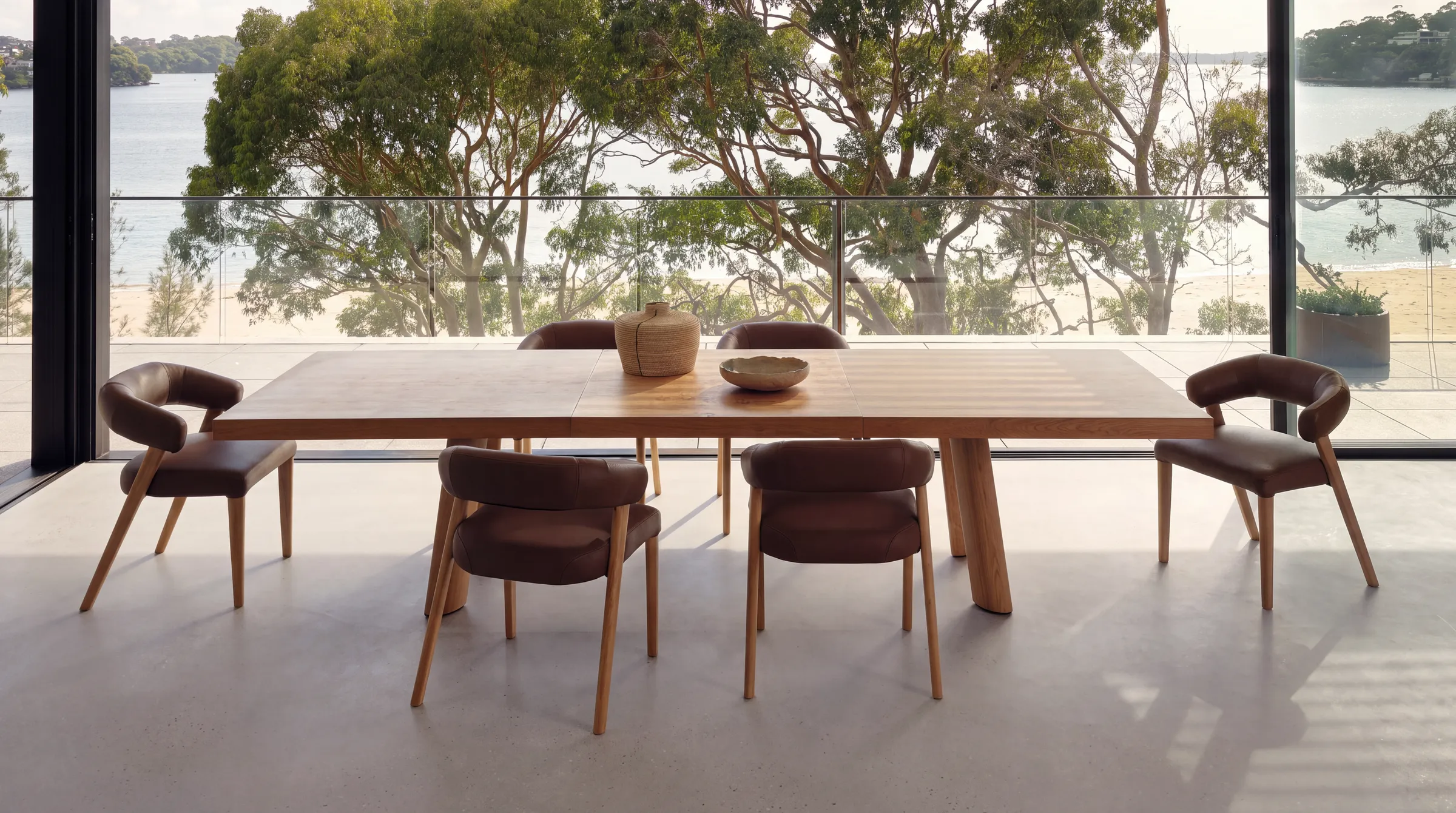 A solid oak dining table with six curved back chairs in wood and brown leather beside a full height glass wall overlooking water and trees.