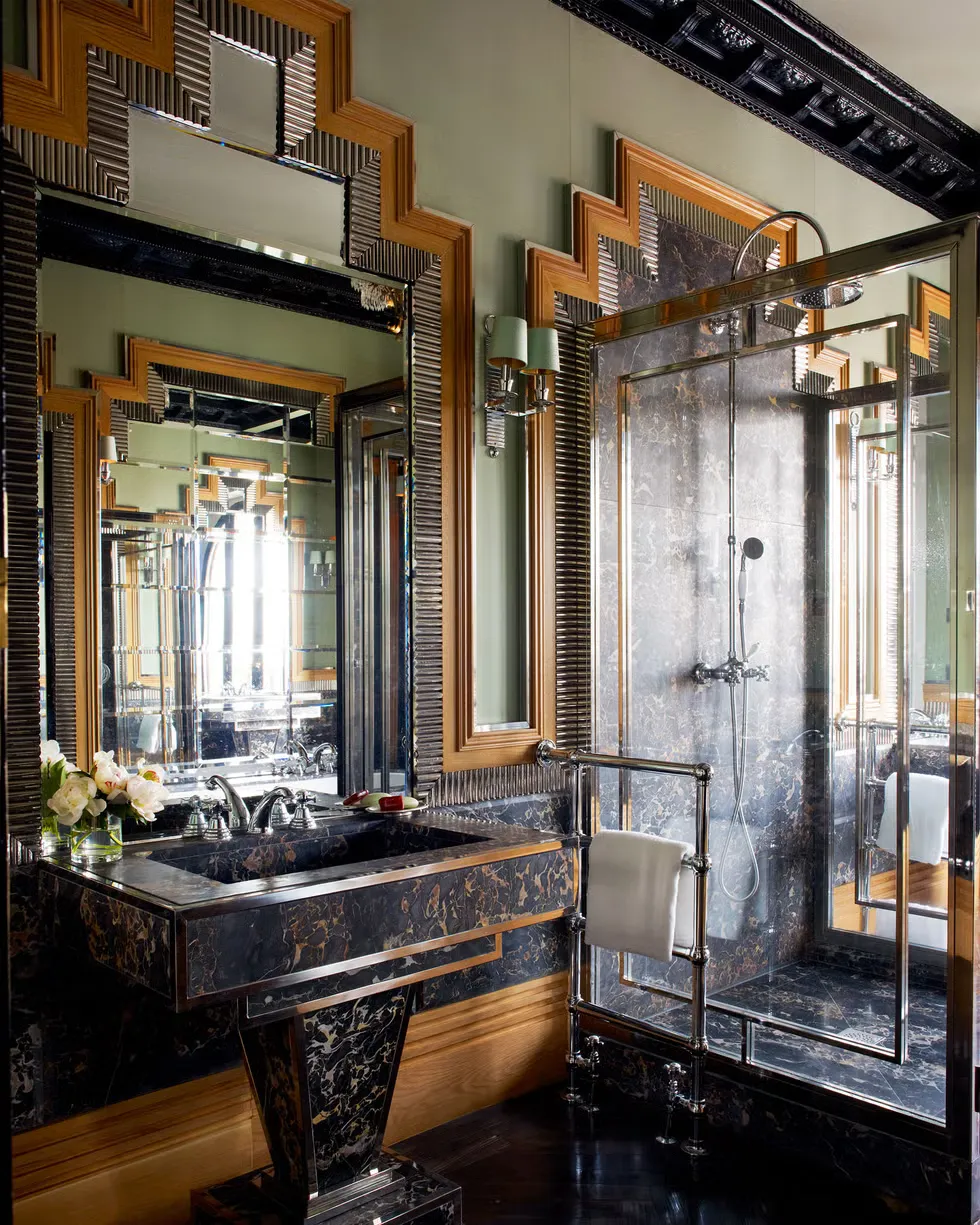 Art Deco bathroom with stepped mirrored frames, a black and gold marble washstand and chrome fittings against sage green walls.