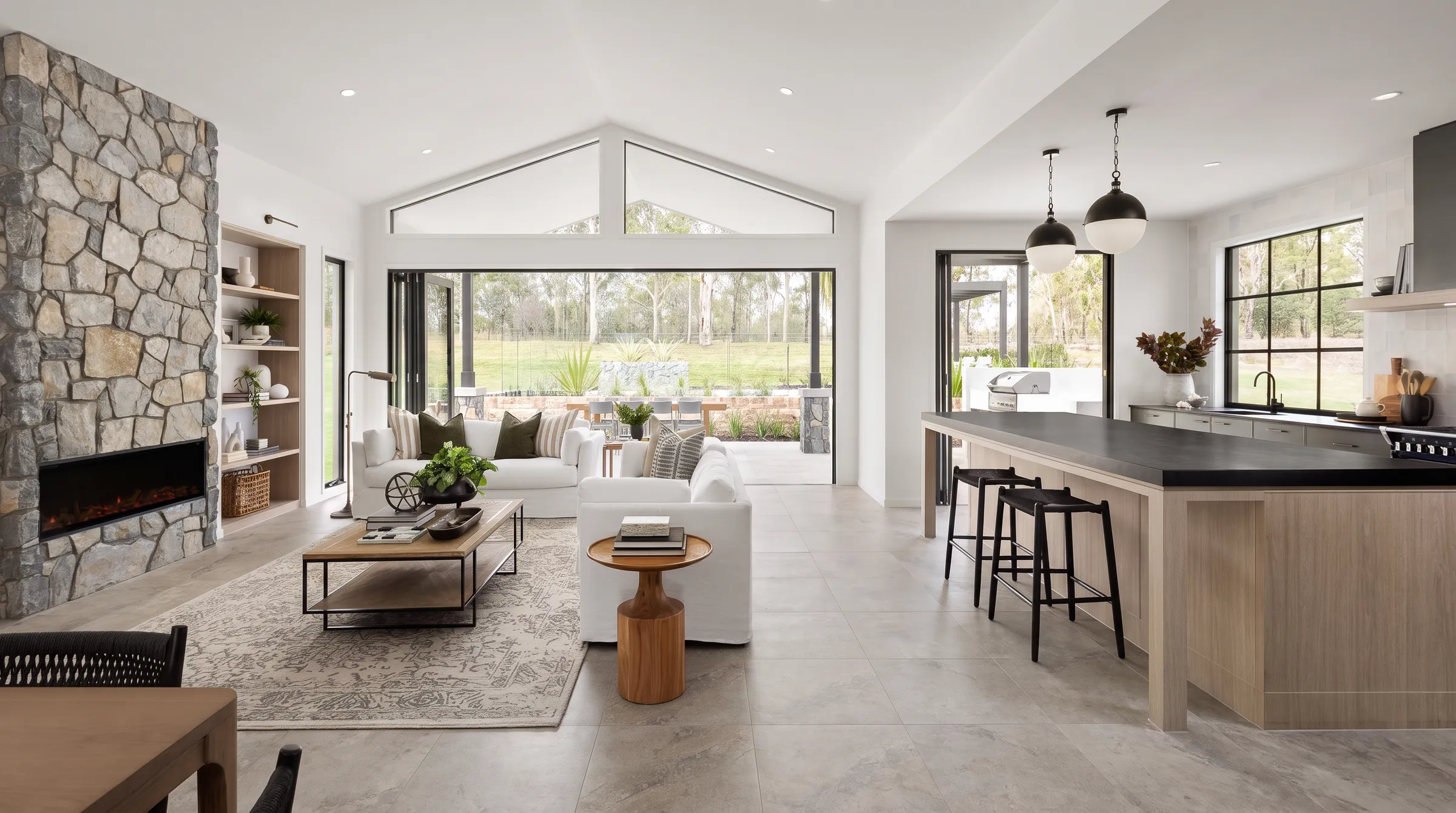 Open plan modern farmhouse living room and kitchen with a stone fireplace, black framed windows, a vaulted ceiling and black globe pendants over a pale oak island.