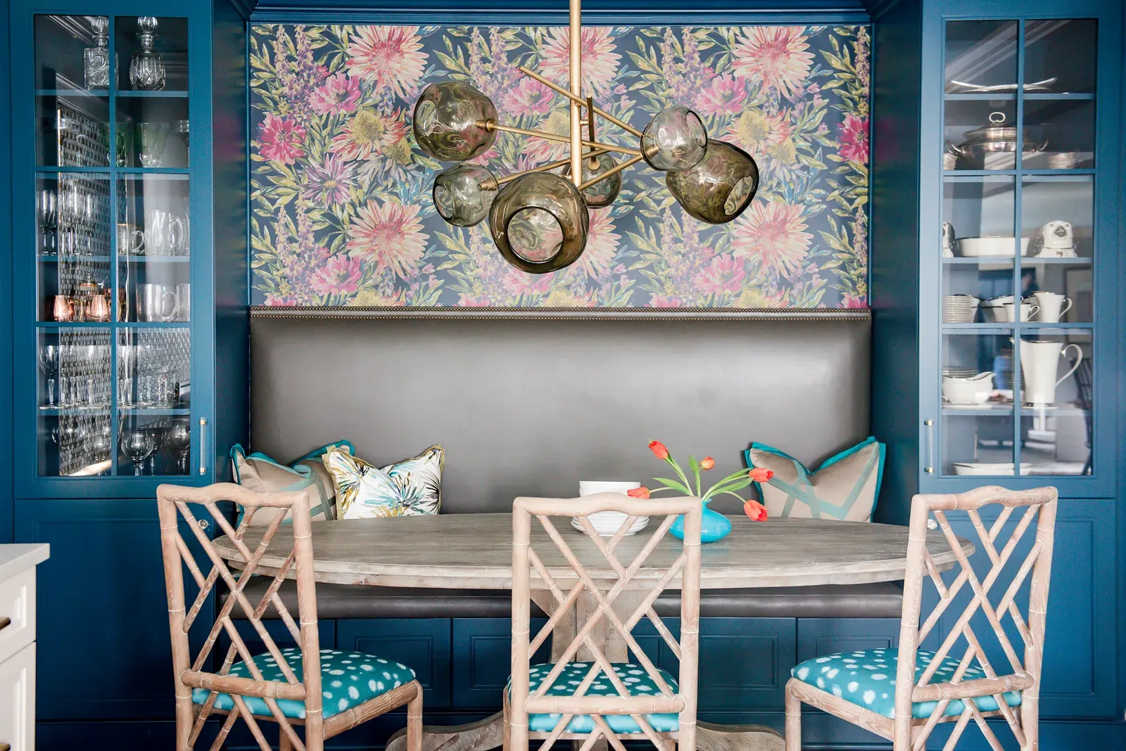A dining nook built into blue cabinetry, with floral wallpaper above a grey leather banquette and glass fronted display cupboards on both sides.