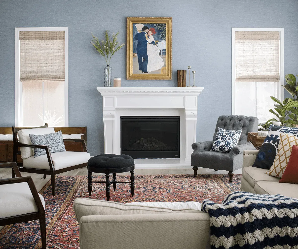 Living room with a classic white fireplace surround and cornice, blue grey walls, a tufted armchair, two slim timber framed chairs and a worn patterned rug.