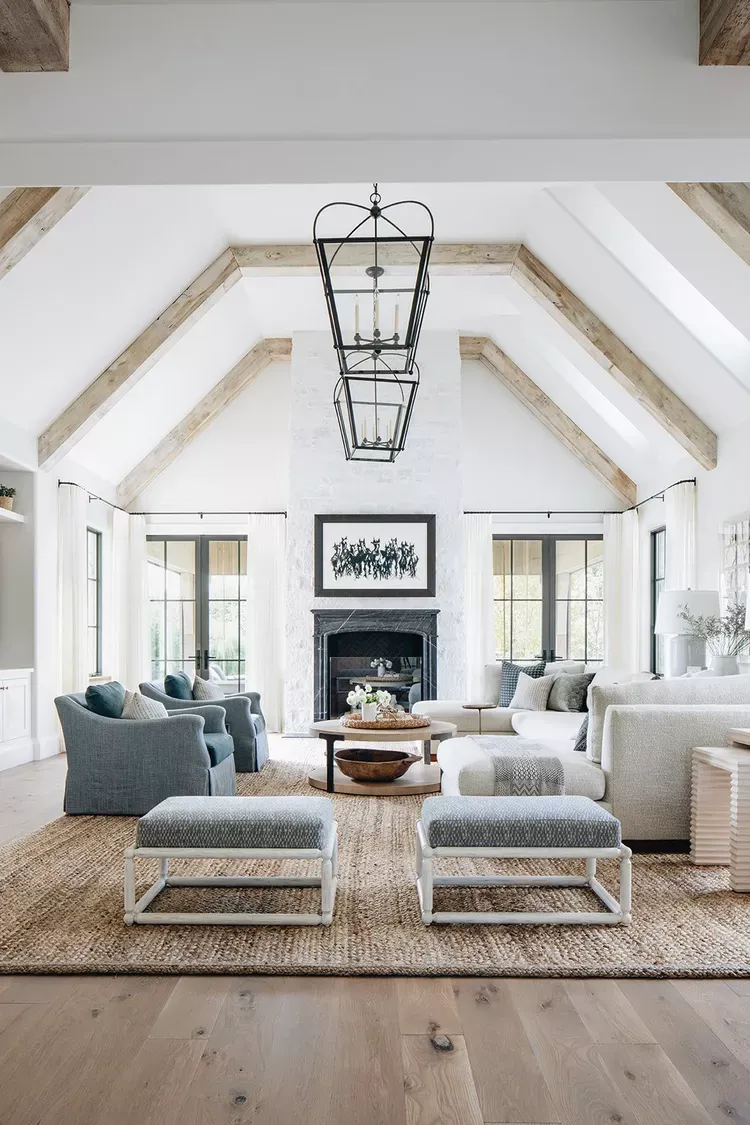 Symmetrical living room with two facing blue armchairs, a pair of matching upholstered benches, a jute rug and a stone fireplace under exposed timber beams.