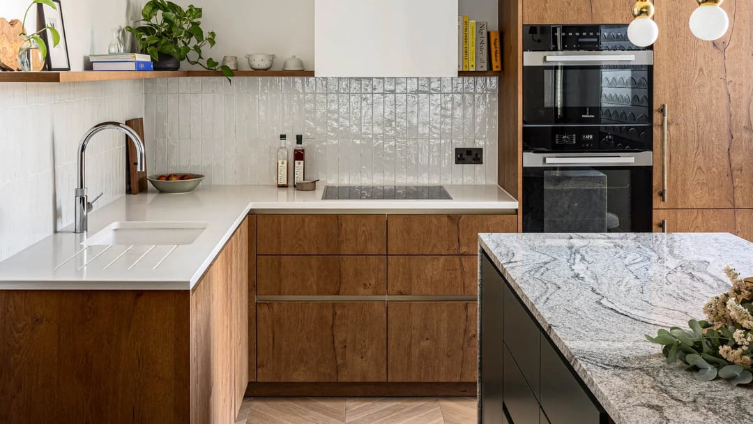 Modern L-shaped kitchen with handleless warm oak fronts, a white worktop, a black island with grey stone and brass pendants.