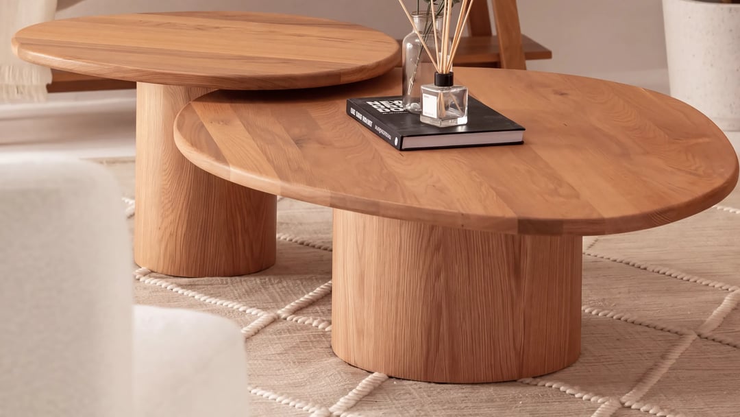 Two nesting oval oak coffee tables on round pedestals, one under the other, on a beige rug with a book and a diffuser on top