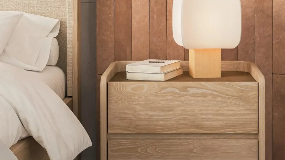 Two-drawer oak nightstand beside a bed, its top level with the mattress, a white lamp on it