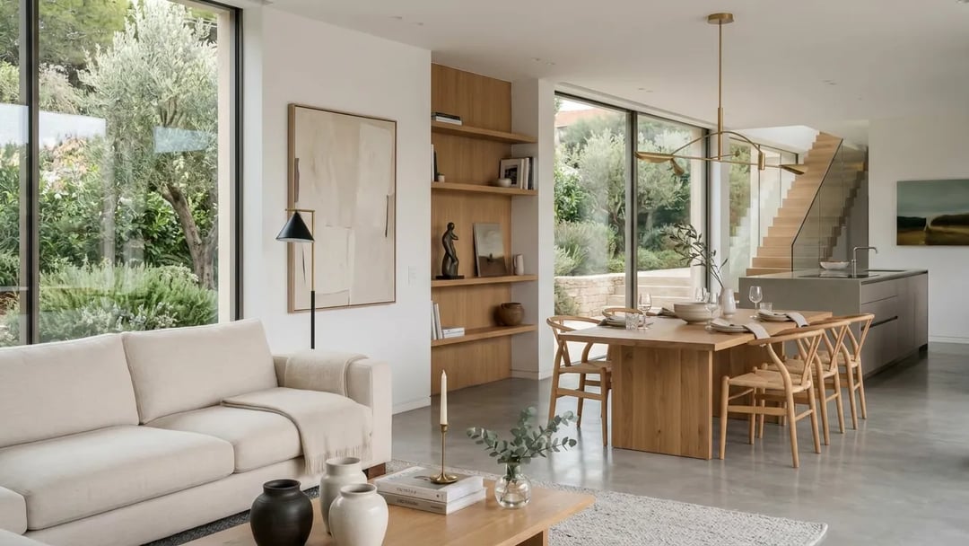 Open plan living and dining room with a cream sofa, an oak dining table with woven chairs under a brass pendant, and garden views through full height glazing.