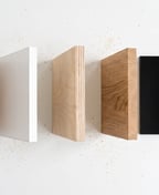 Five furniture board samples against a white wall: bare MDF, white melamine, birch plywood, solid oak and black acrylic.