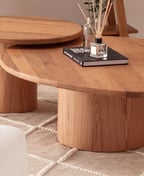 Two nesting oval oak coffee tables on round pedestals, one under the other, on a beige rug with a book and a diffuser on top