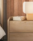 Two-drawer oak nightstand beside a bed, its top level with the mattress, a white lamp on it