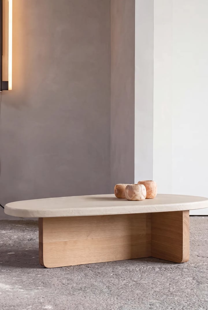 Oval coffee table with a travertine top on a cross-shaped oak base, two small marble bowls on top, against a grey wall