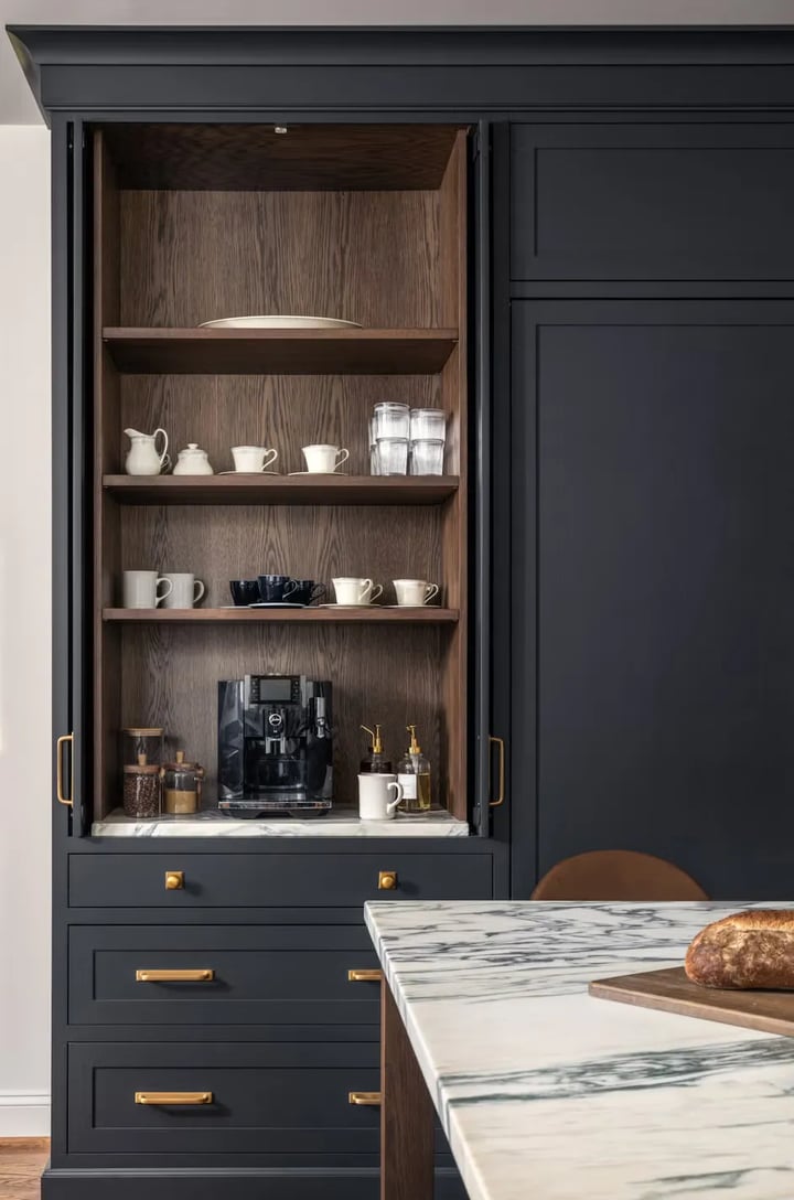 Dark blue tall pantry unit with pocket doors open on oak-lined shelves, a coffee machine and cups, brass handles.