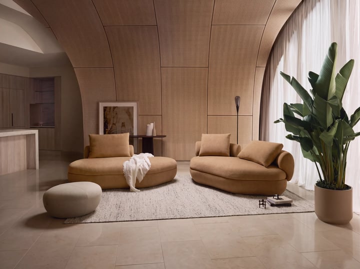 Living room with a curved perforated wood panel wall, two rounded camel sofas, a pebble shaped ottoman and a cream rug.