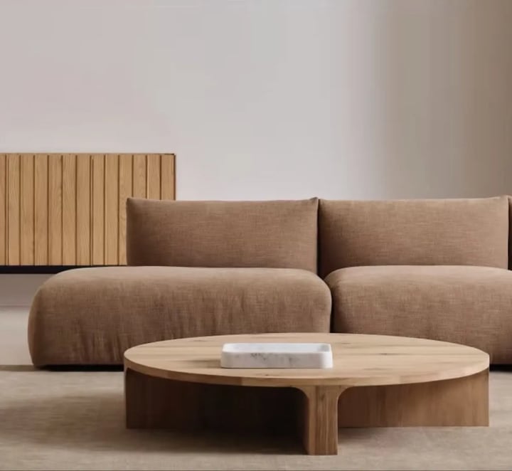 Round oak coffee table with a marble tray in front of a low camel modular sofa, on a beige rug