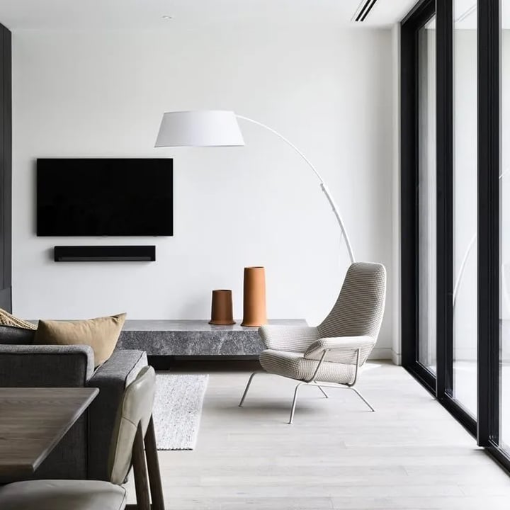 A grey sofa and a woven lounge chair beside a low stone bench holding two terracotta vessels, lit by a white arc floor lamp.