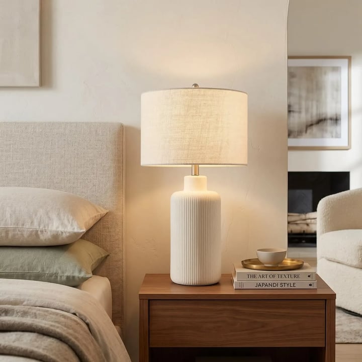 Walnut nightstand with a white ceramic lamp on it, books and a bowl, beige upholstered headboard