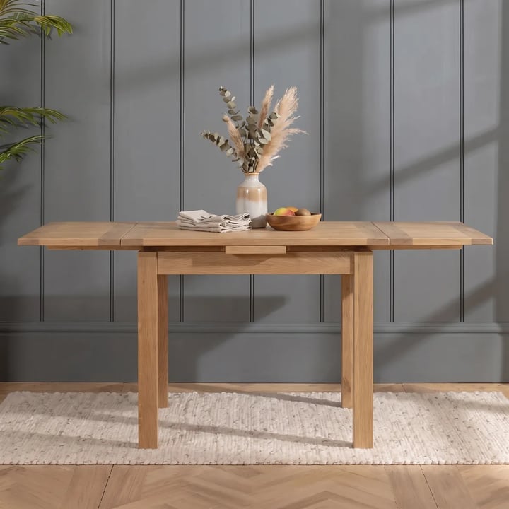 Small oak dining table with its long side against a grey panelled wall and the leaves pulled out at both ends, on a rug
