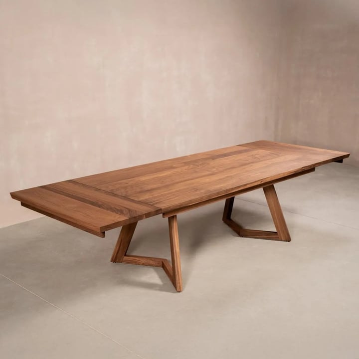 Long walnut dining table with extension leaves at both ends on two angular wooden supports, in an empty room