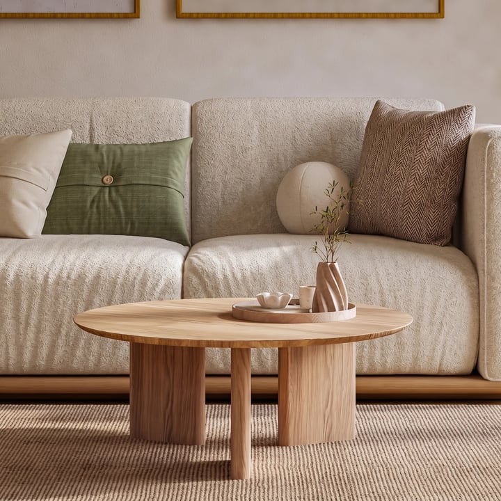 Round oak coffee table on three curved legs in front of a cream boucle sofa with green cushions, on a jute rug
