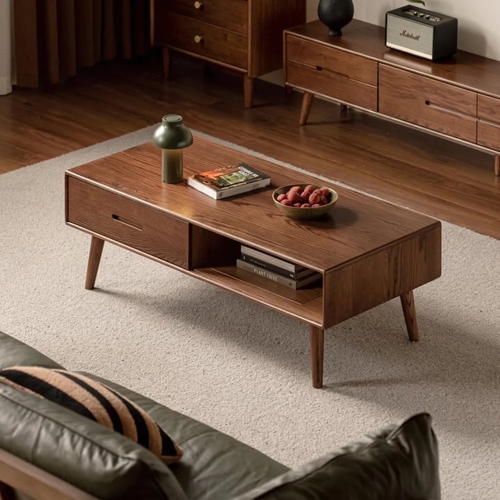 Classic walnut coffee table with a drawer, an open shelf and splayed legs on a cream rug, a green leather sofa in front