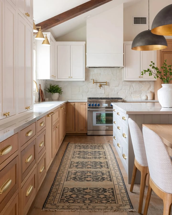 Kitchen with cream wall cabinets, light oak base units with brass cup handles, a greige island and a white stone wall.