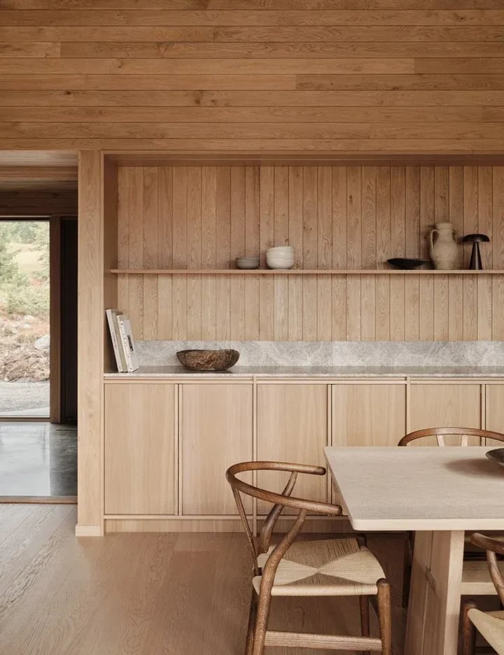 Oak kitchen run with a pale stone worktop and an open timber shelf above it, beside a dining table with chairs.