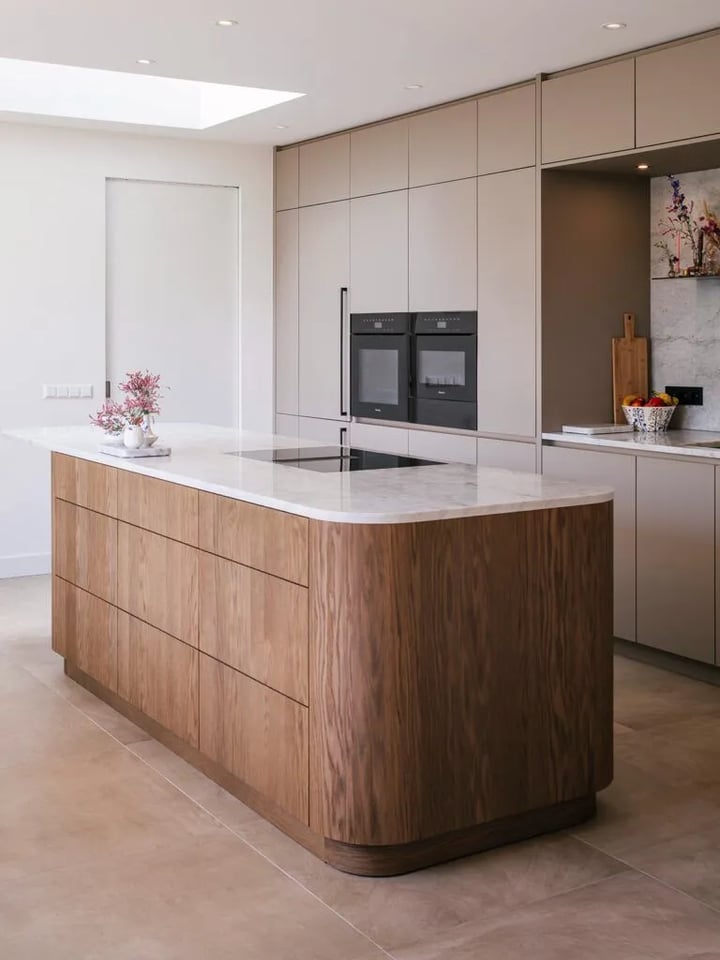 Curved walnut island with a white marble worktop and hob, with handleless greige tall units behind it.