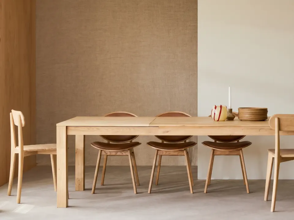 Oak extending dining table open with its centre leaf in place, six oak chairs with leather seats against a textured wall