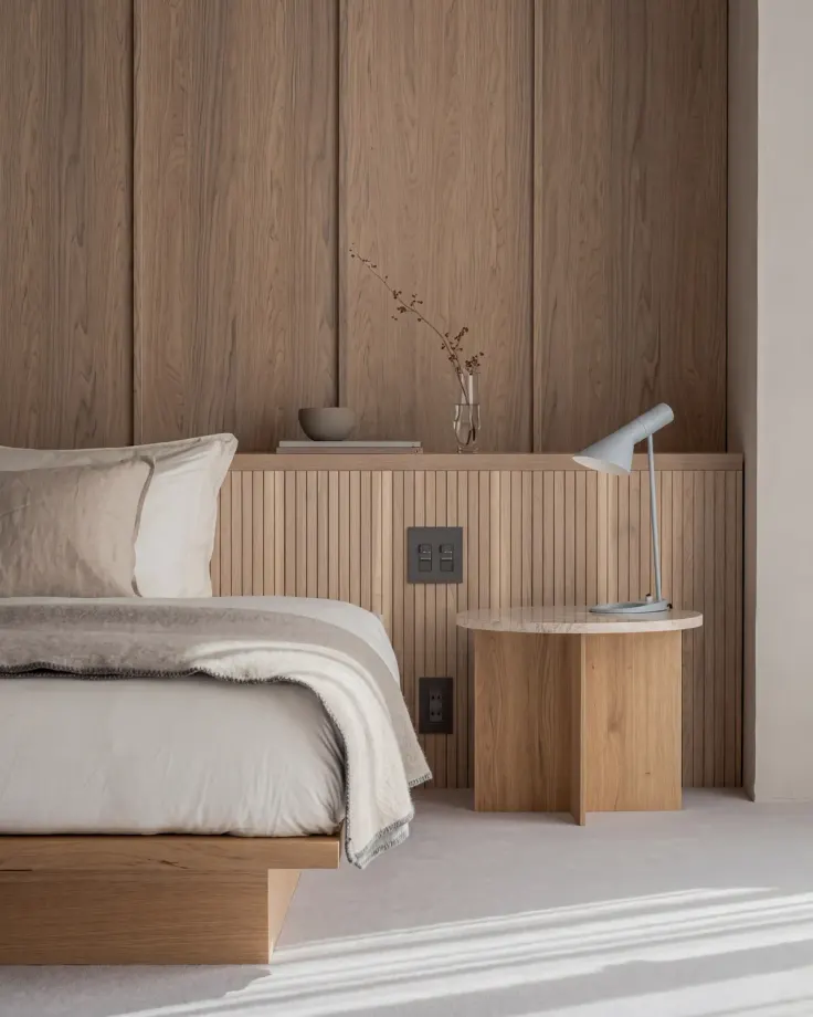 Low oak platform bed against an oak panelled wall with a fluted headboard band, a round oak side table and a lamp