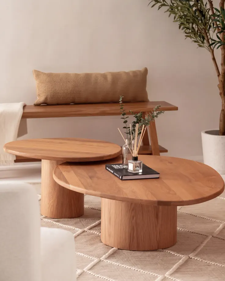 Two nesting oval oak coffee tables on round pedestals in front of an oak bench, a book and a diffuser on top
