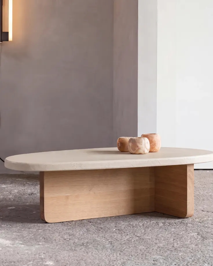 Oval coffee table with a travertine top on an oak plywood base, two small marble bowls on top, against a grey wall